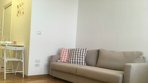 Ein Wohnzimmer mit einem beigen Sofa, zwei karierten Kissen, einem weißen Sessel und einem kleinen Tisch mit Kerze und Gläsern.