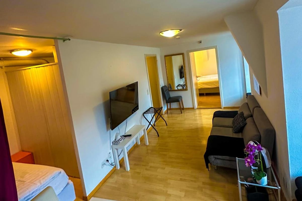 A hotel room with a TV, a sofa, a small table, and a chair. There is a bed with a white headboard and a red curtain.