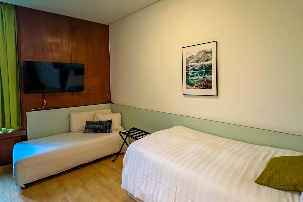 A room with a bed, a sofa, a flat-screen TV mounted on a wooden panel, and a framed picture on the wall.