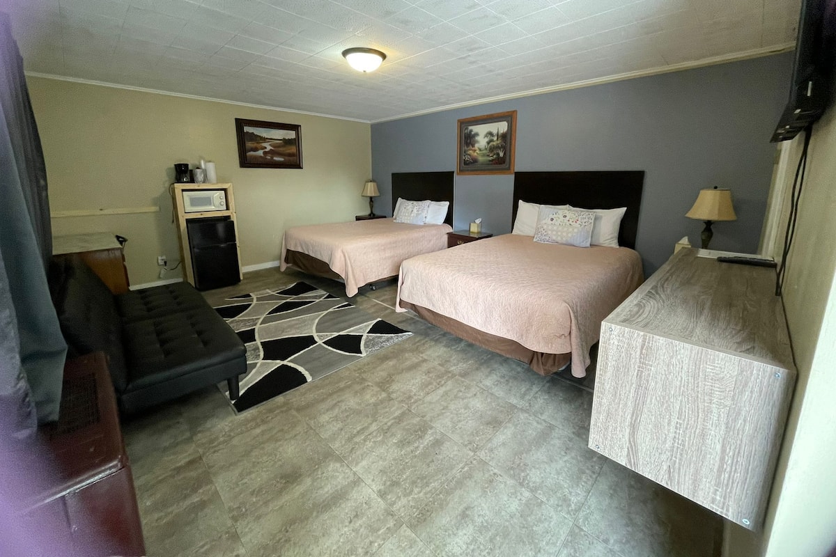 A hotel room with two beds, a desk, a chair, a small kitchenette, and a ceiling light.