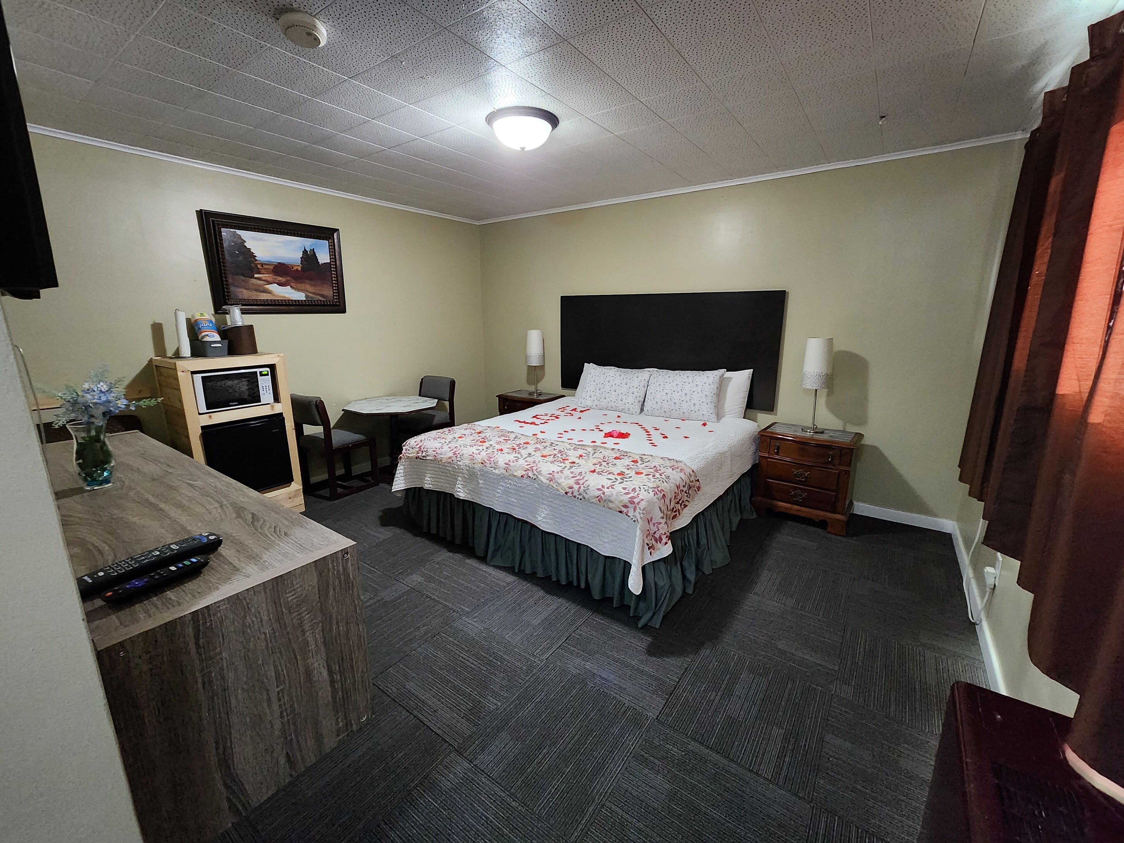 Ein Hotelzimmer mit Bett, Nachttischen, einem Schreibtisch mit Stuhl, einer Mikrowelle, einer Vase mit Blumen und einem Bild an der Wand.
