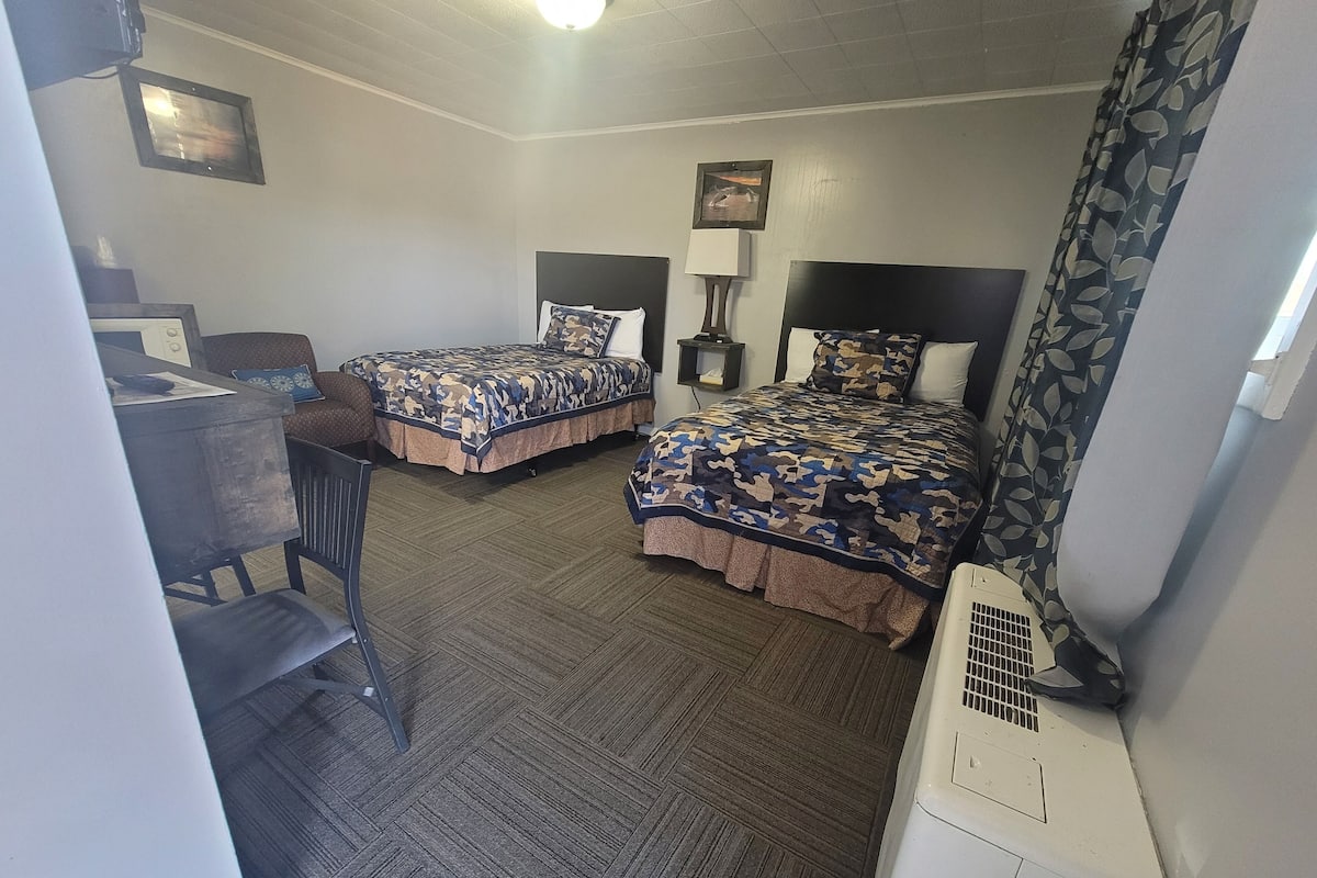 A hotel room with two beds, a desk, a chair, a heater, and a window with curtains.