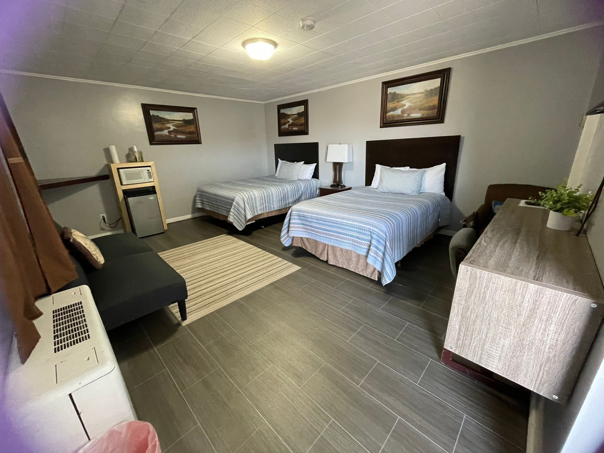 A hotel room with two beds, a desk, a chair, a small table, a lamp, a plant, and a picture frame on the wall.