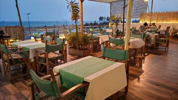 Un restaurant en plein air avec des tables et des chaises en bois, une vue sur la plage et un toit de chaume.