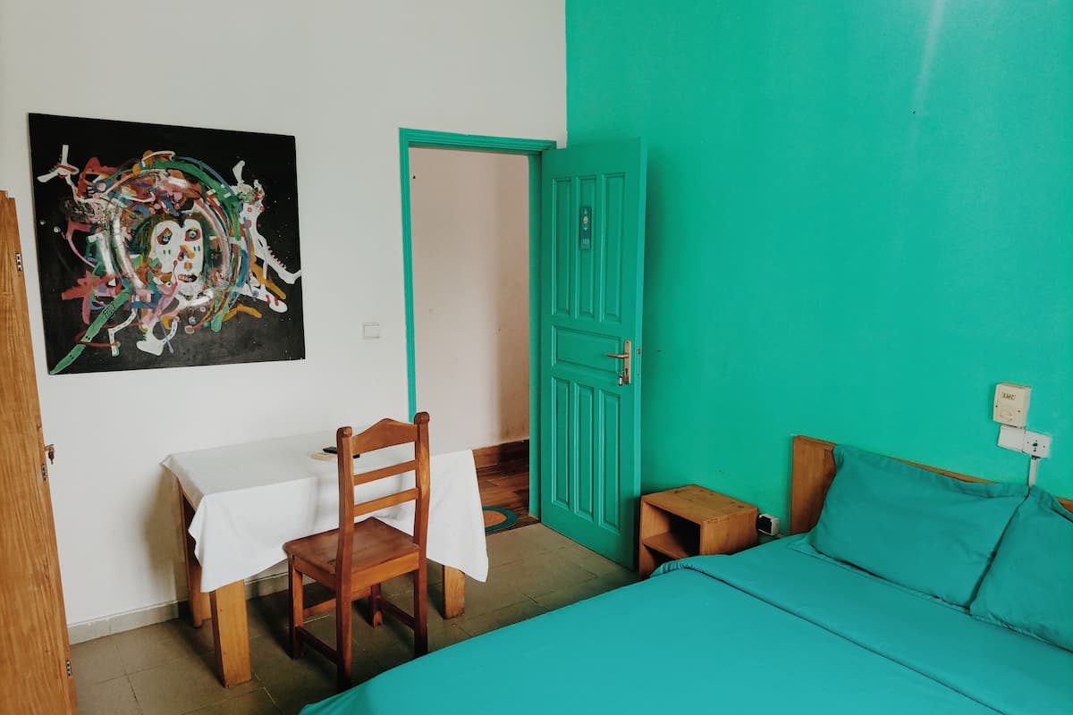 A room with a bed, a table with a chair, a door, and a wall with a colorful painting.