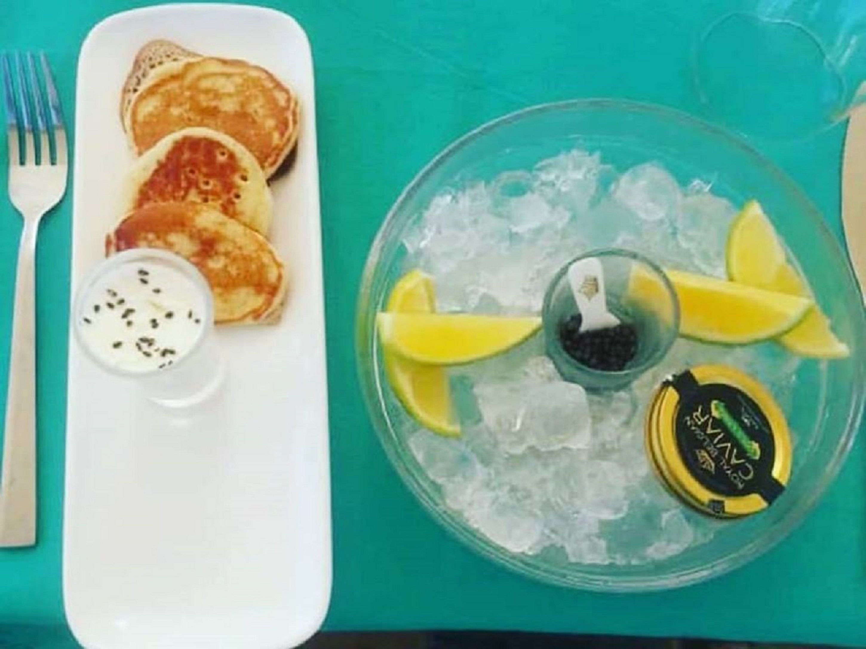 A plate of pancakes with a side of yogurt and a glass dish with lemon slices and a bottle of sauce.