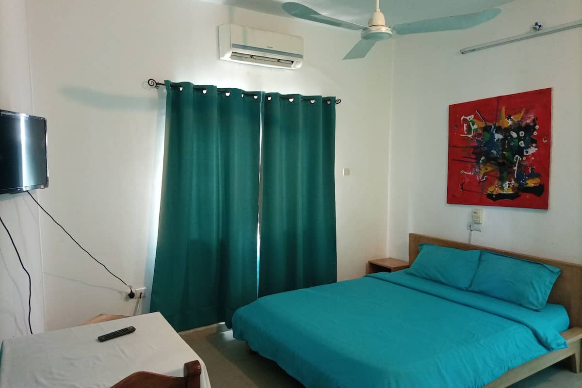 A bedroom with a bed, a desk, a chair, an air conditioner, a ceiling fan, and a painting on the wall.