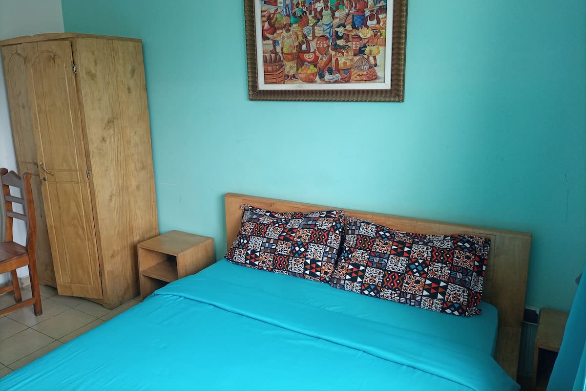 A bedroom with a bed, a wooden wardrobe, a small bedside table, and a colorful framed picture on the wall.
