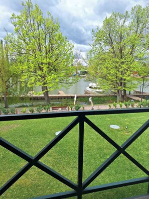 A view from a balcony with a grassy lawn, a wooden dock, and a body of water with boats. - Duna Garden Hotel (Budapest)