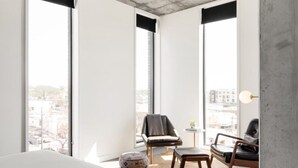 Ein modernes Schlafzimmer mit einem großen Bett, zwei Sessel, einem kleinen Tisch und Blick auf die Stadt durch große Fenster.