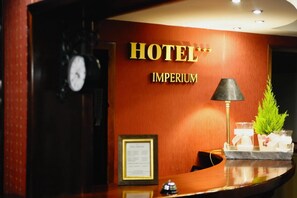 A hotel reception desk with a clock, a framed document, and a plant.
