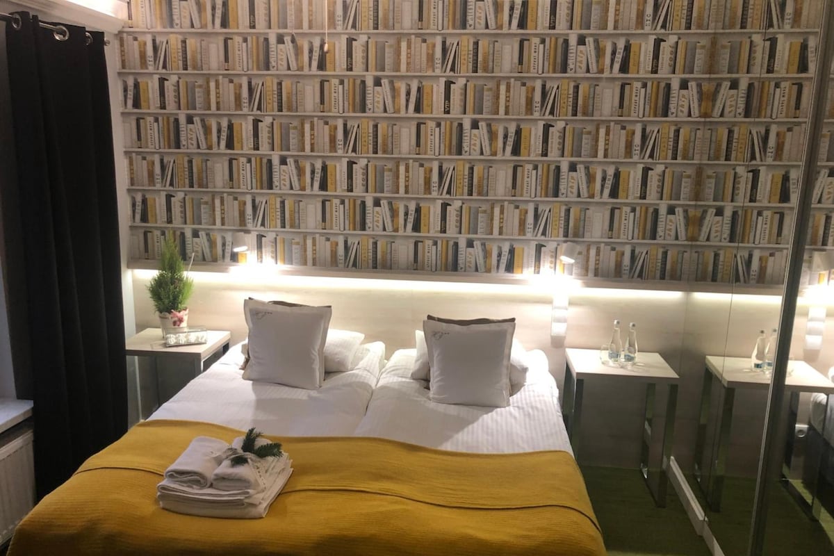 Hotellihuone, jossa on suuri kirjahylly täynnä kirjoja, valkoisilla lakanoilla varustettu sänky sekä pieni pöytä, jonka päällä on kasvi ja pullo.