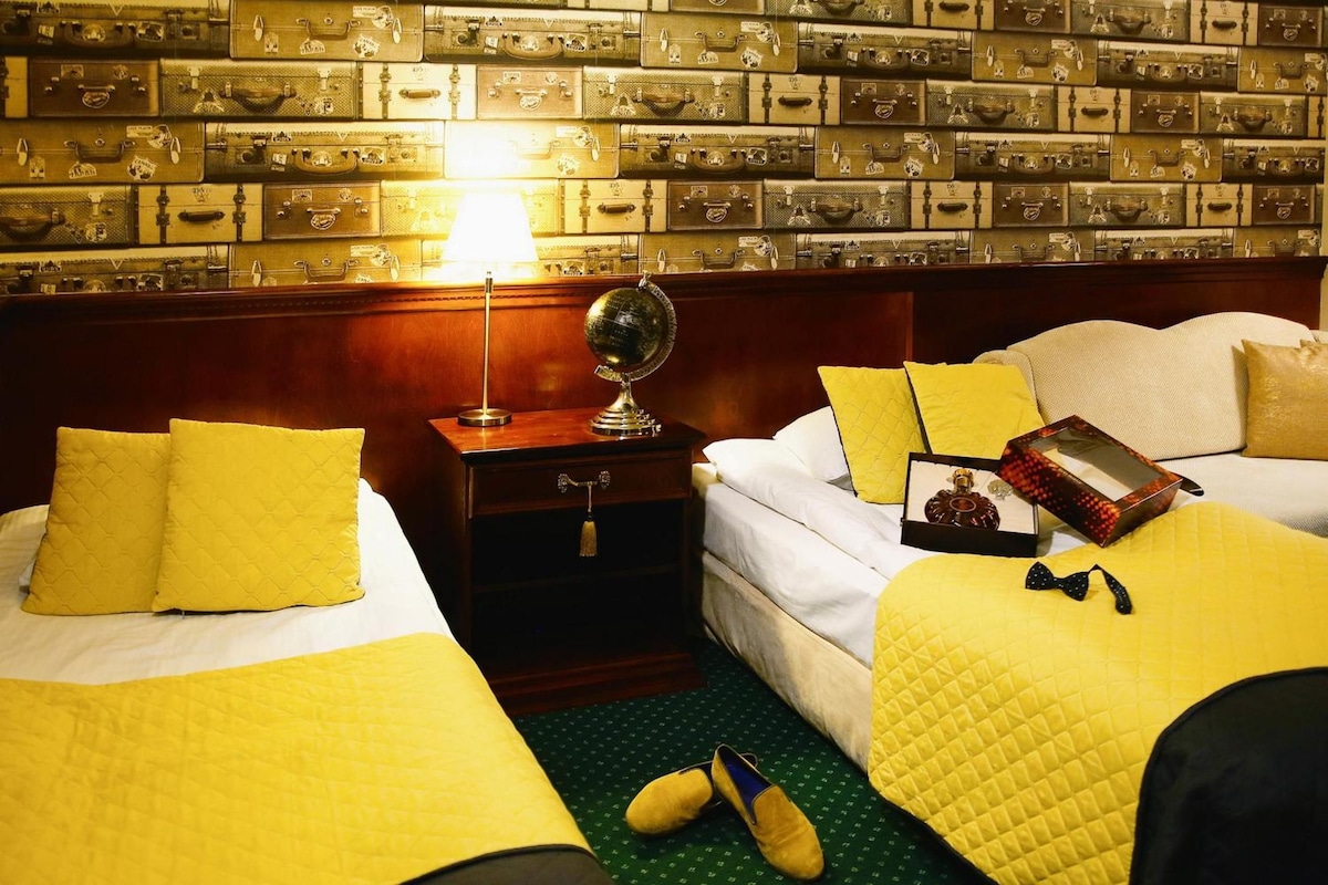 A hotel room with two beds, a nightstand, a lamp, and a wall of suitcases.