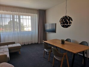 Habitación de hotel con un ventanal grande, televisor de pantalla plana, mesa de madera con sillas, sofá y una lámpara esférica original.