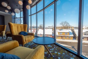 Un hall de hotel con grandes ventanales, un sillón amarillo, una mesa redonda y una almohada azul.