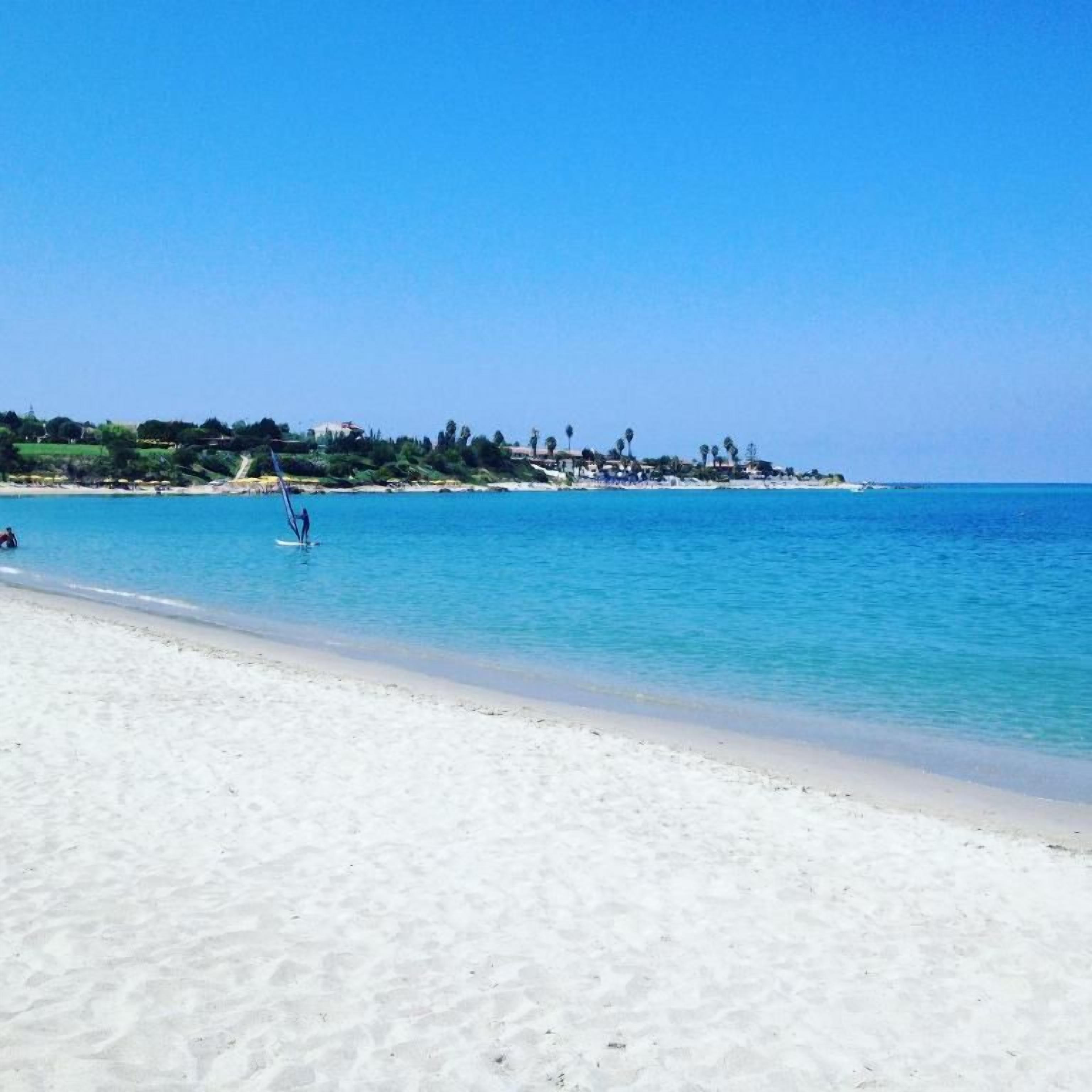 Una playa con agua azul cristalina, una orilla arenosa y una costa lejana con edificios.