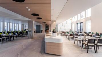 A spacious, modern dining area with multiple tables and chairs, a central counter, and large windows allowing natural light.