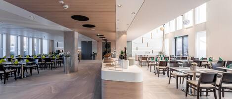 Un comedor amplio y moderno con múltiples mesas y sillas, una barra central y ventanas grandes que dejan entrar luz natural.