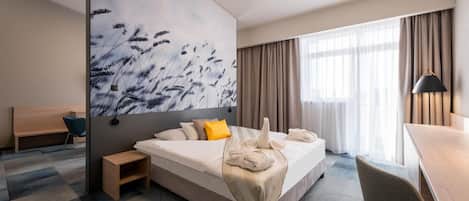 A modern hotel room with a large bed, a desk, a chair, and a painting on the wall.