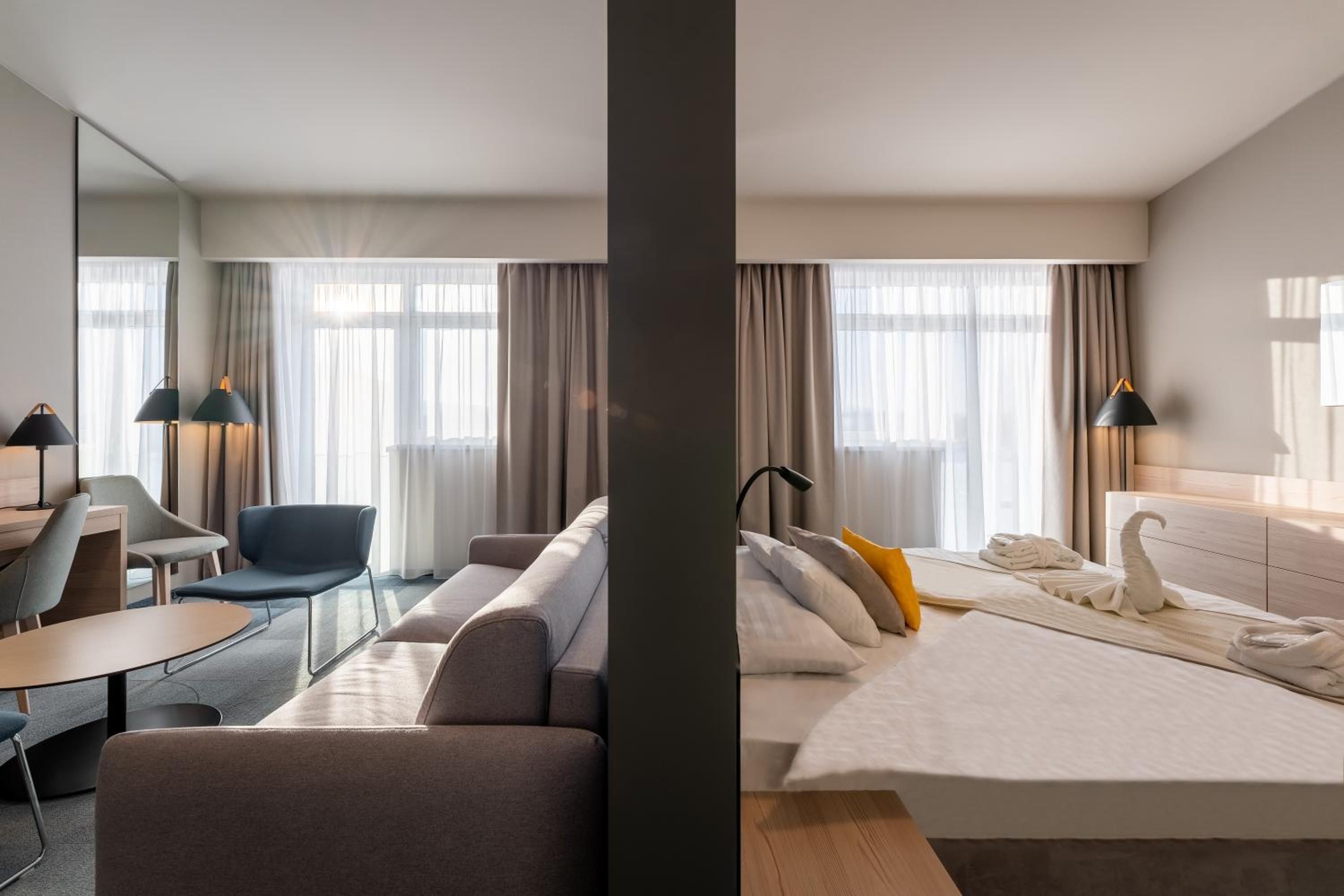 A hotel room with a sofa, a desk, a chair, a bed with a white duvet, and a large mirror.