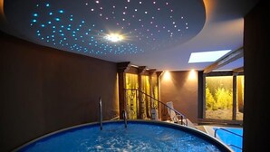 Un jacuzzi con techo que proyecta luces simulando estrellas, vista de un bosque a través de un ventanal grande y una piscina contigua al jacuzzi.