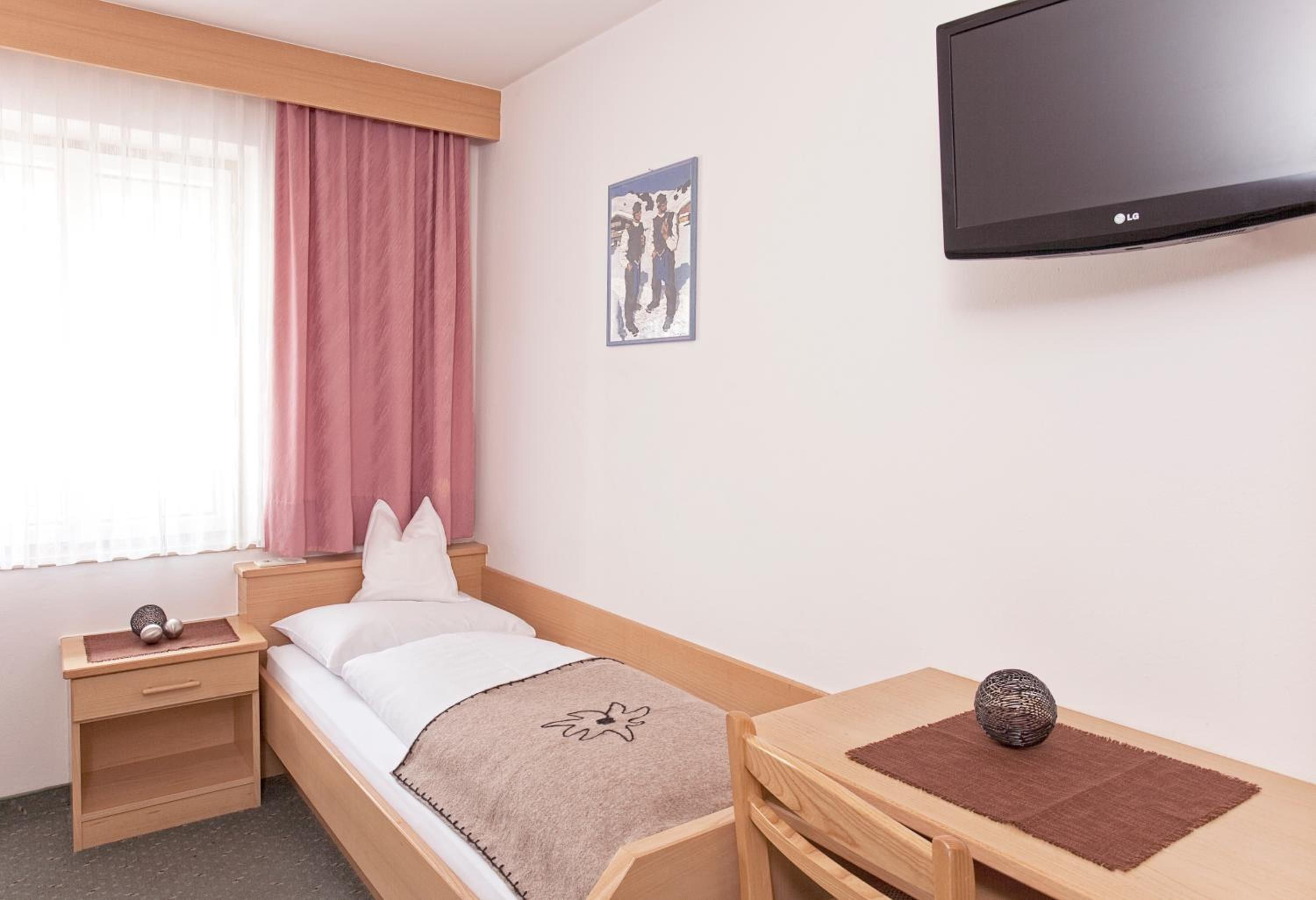 Une chambre d’hôtel avec un lit simple, une table de chevet, un petit bureau avec une chaise, un téléviseur fixé au mur et une fenêtre avec des rideaux.