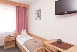 Una habitación de hotel con una cama individual, una mesita de noche, un escritorio pequeño con una silla, un televisor montado en la pared y una ventana con cortinas.