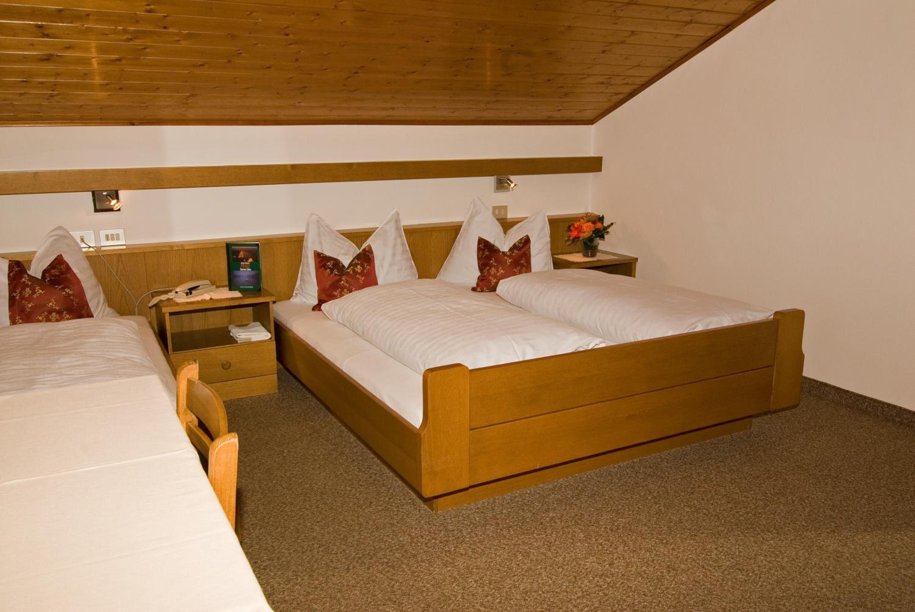 Ein Hotelzimmer mit einem hölzernen Bett, einem Nachttisch mit Lampe sowie einem weiteren Tisch mit Telefon und Buch.