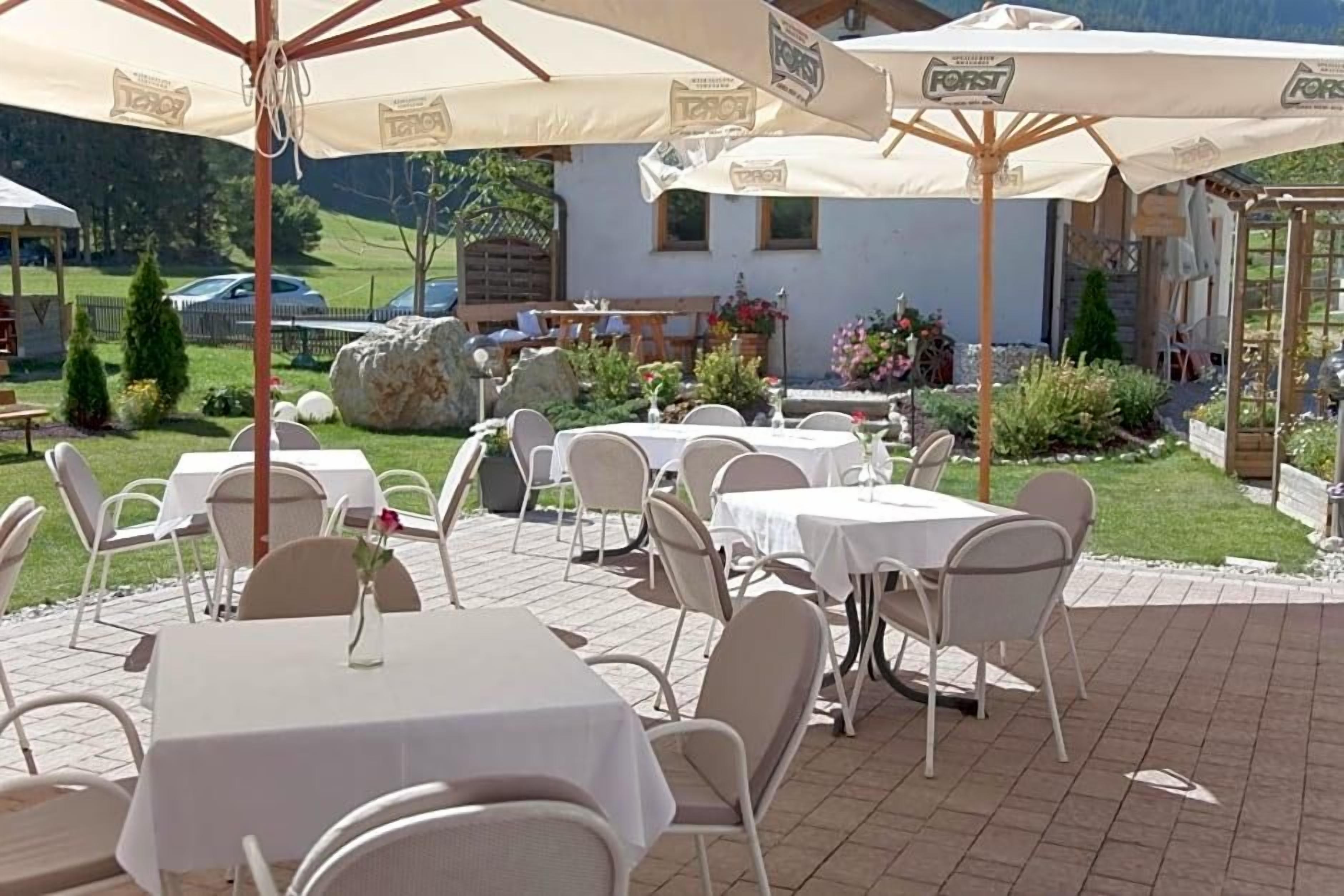 An outdoor patio area with tables and chairs, some covered by umbrellas, and a view of a building and greenery in the background.
