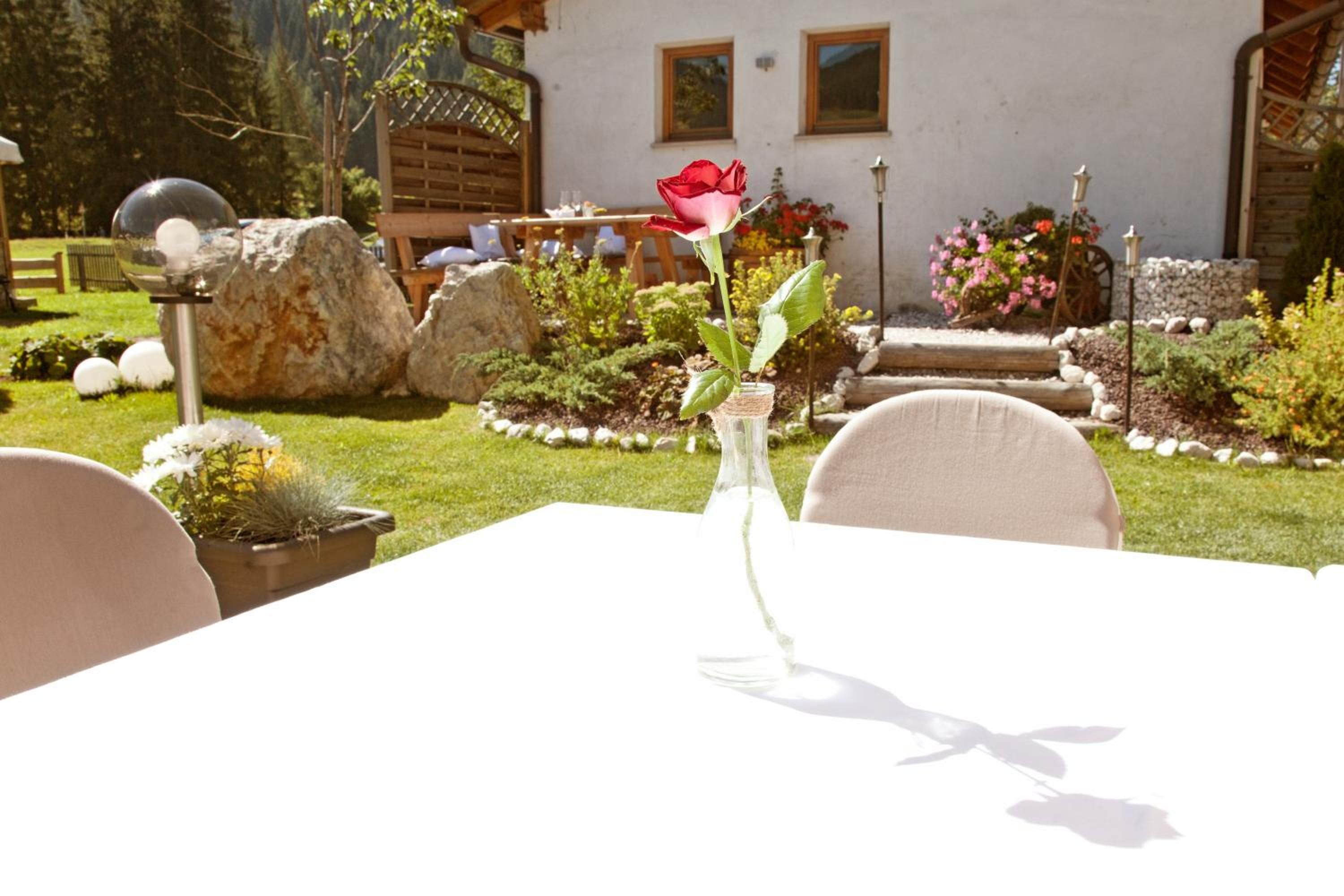 Uma mesa com um vaso contendo uma única rosa, em um jardim com um edifício branco ao fundo.