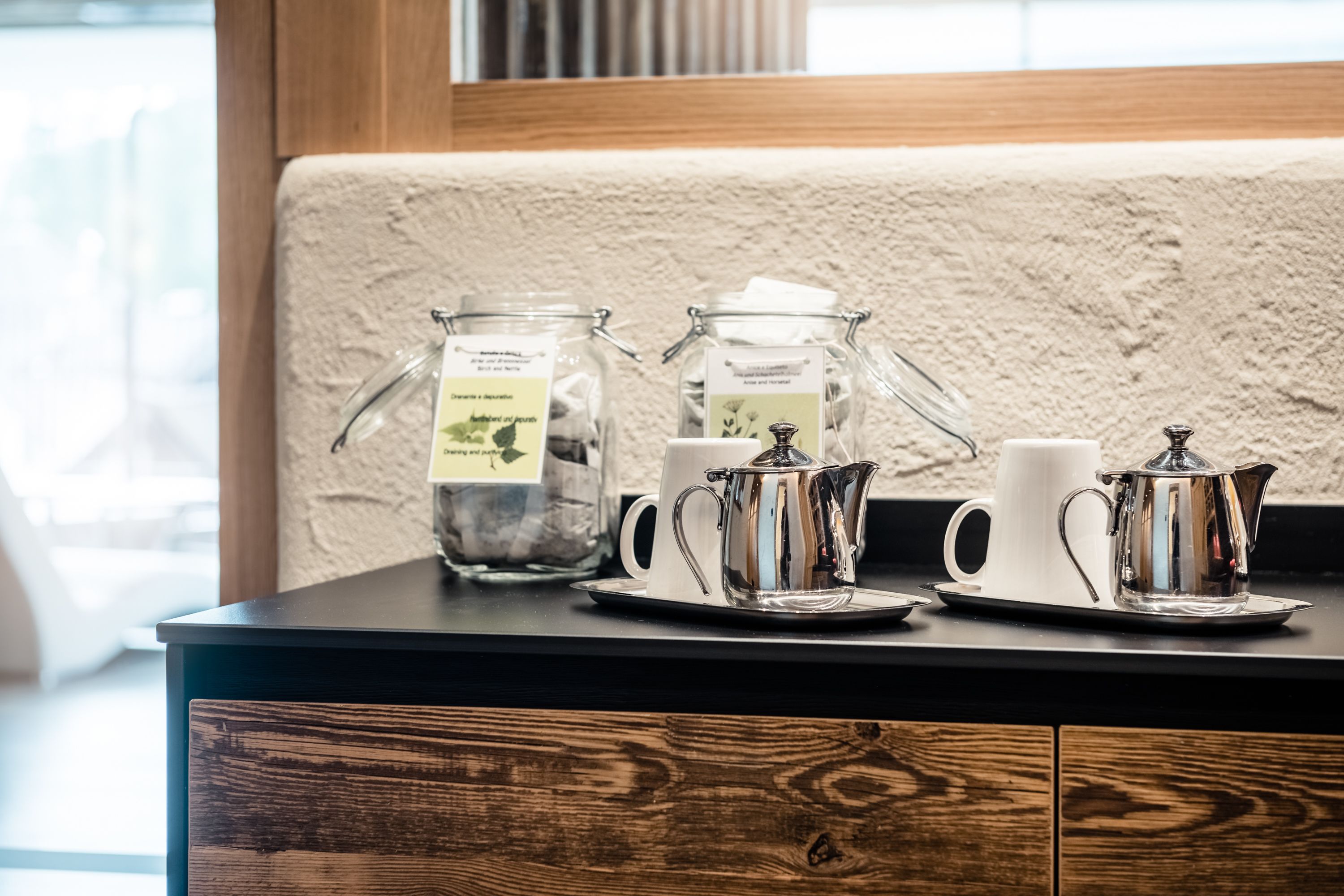 A coffee station with a teapot, cups, and jars of tea on a wooden counter.