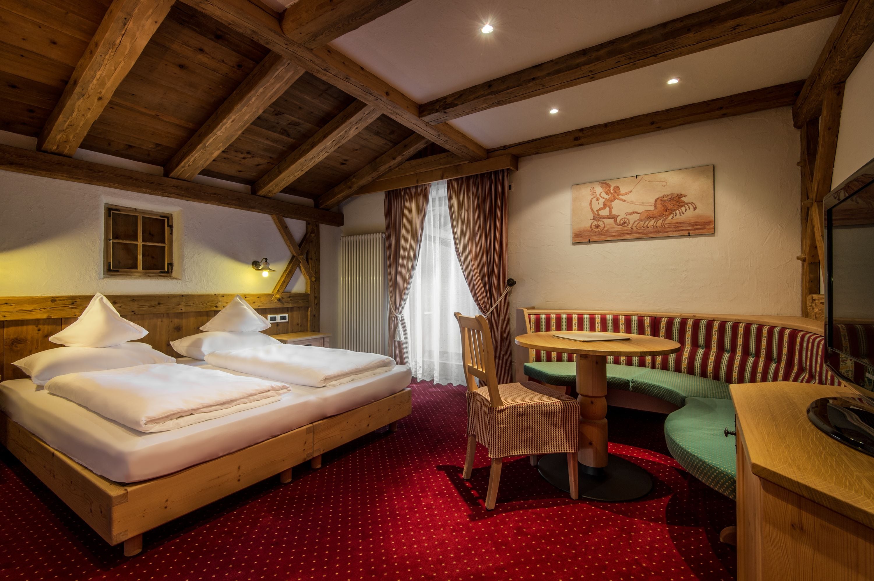 A room with a wooden ceiling, a bed with white linens, a green upholstered bench, a small round table, and a painting on the wall.