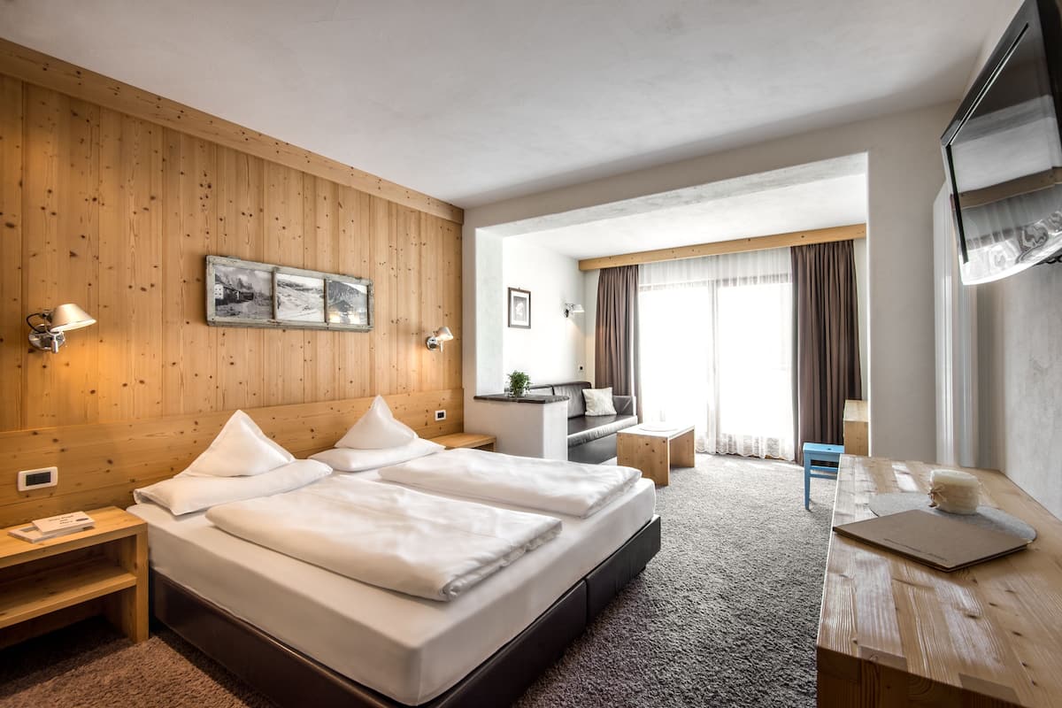 A hotel room with a wooden wall, a bed with white linens, a desk, a chair, and a window with curtains.