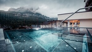 A modern outdoor pool area with clear glass flooring, surrounded by a metal railing, and overlooking a mountainous landscape. - Hotel Gran Ciasa (Corvara in Badia)