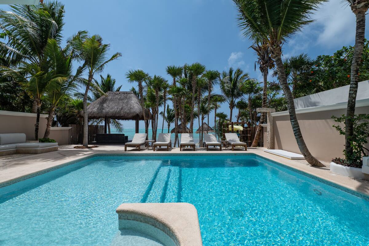 A pool area with lounge chairs, palm trees, and a thatched-roof structure.