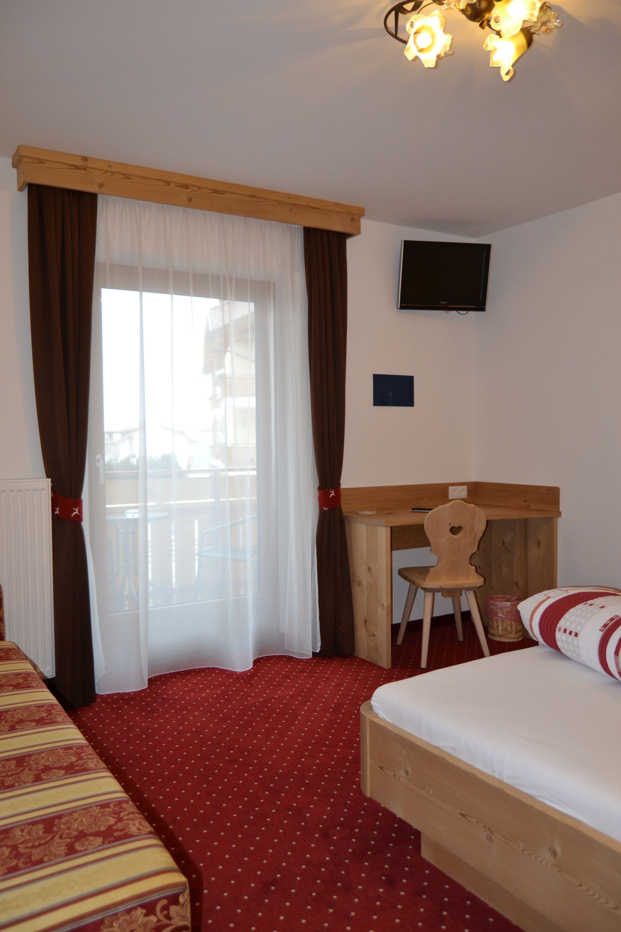 Une chambre d’hôtel avec un lit, un bureau et une chaise. Il y a une fenêtre avec des rideaux, un téléviseur fixé au mur et une moquette rouge.