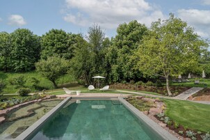 Una piscina rectangular con borde de piedra, rodeada de un césped cuidado y árboles. Se observan sillones de descanso y sombrillas al fondo.