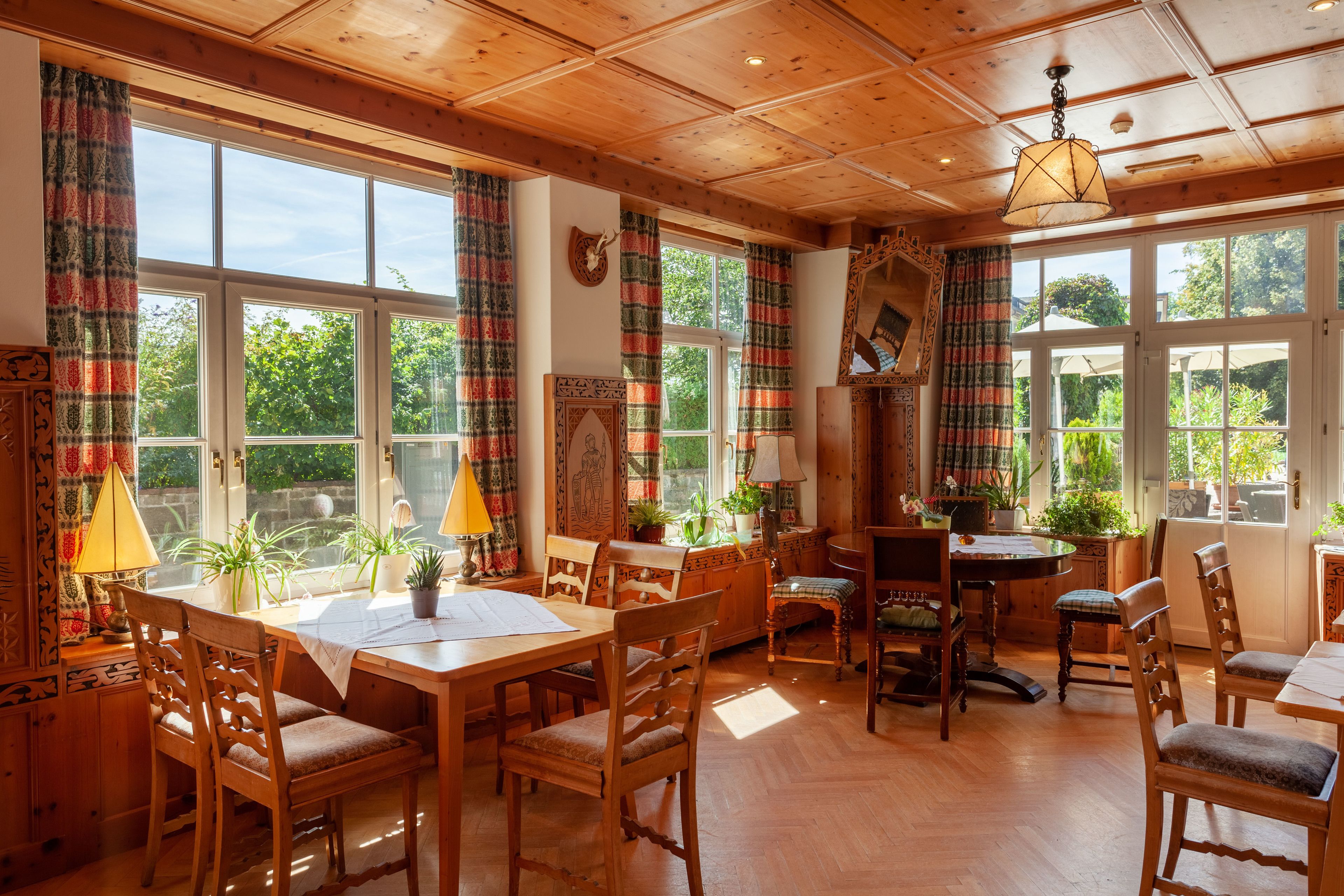 A wooden dining room with a table and chairs, large windows with curtains, and a ceiling with wooden beams.