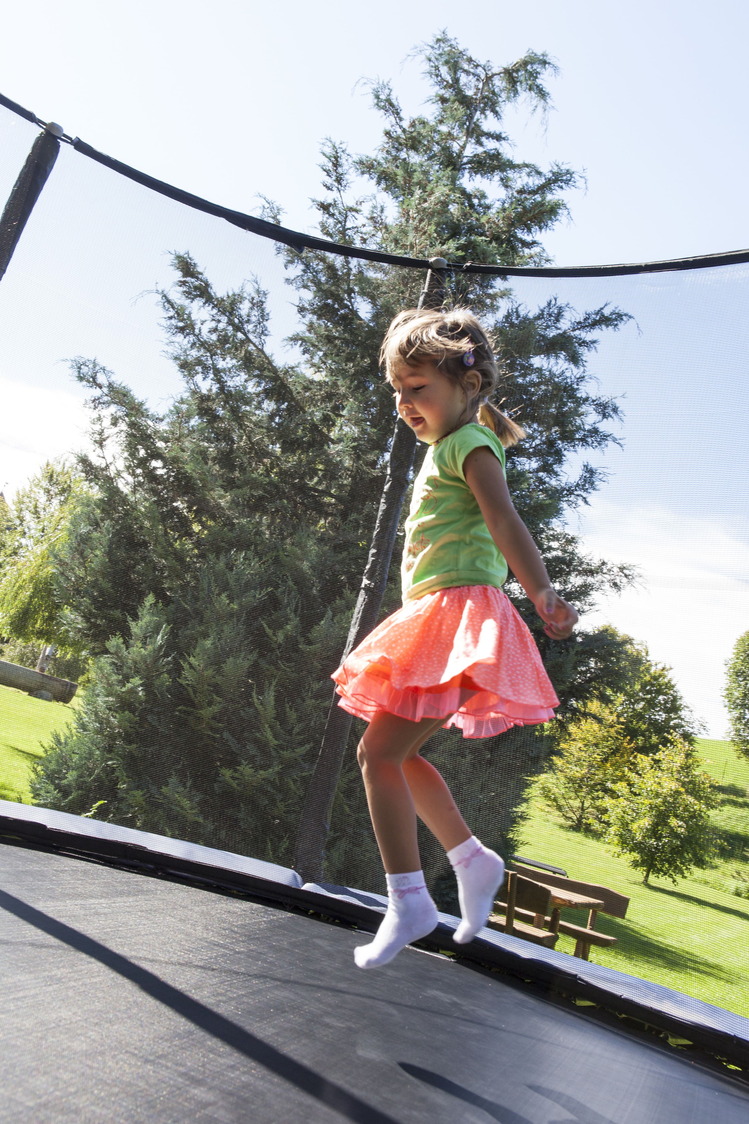 A child on a trampoline with a netted enclosure, surrounded by trees and a grassy area.