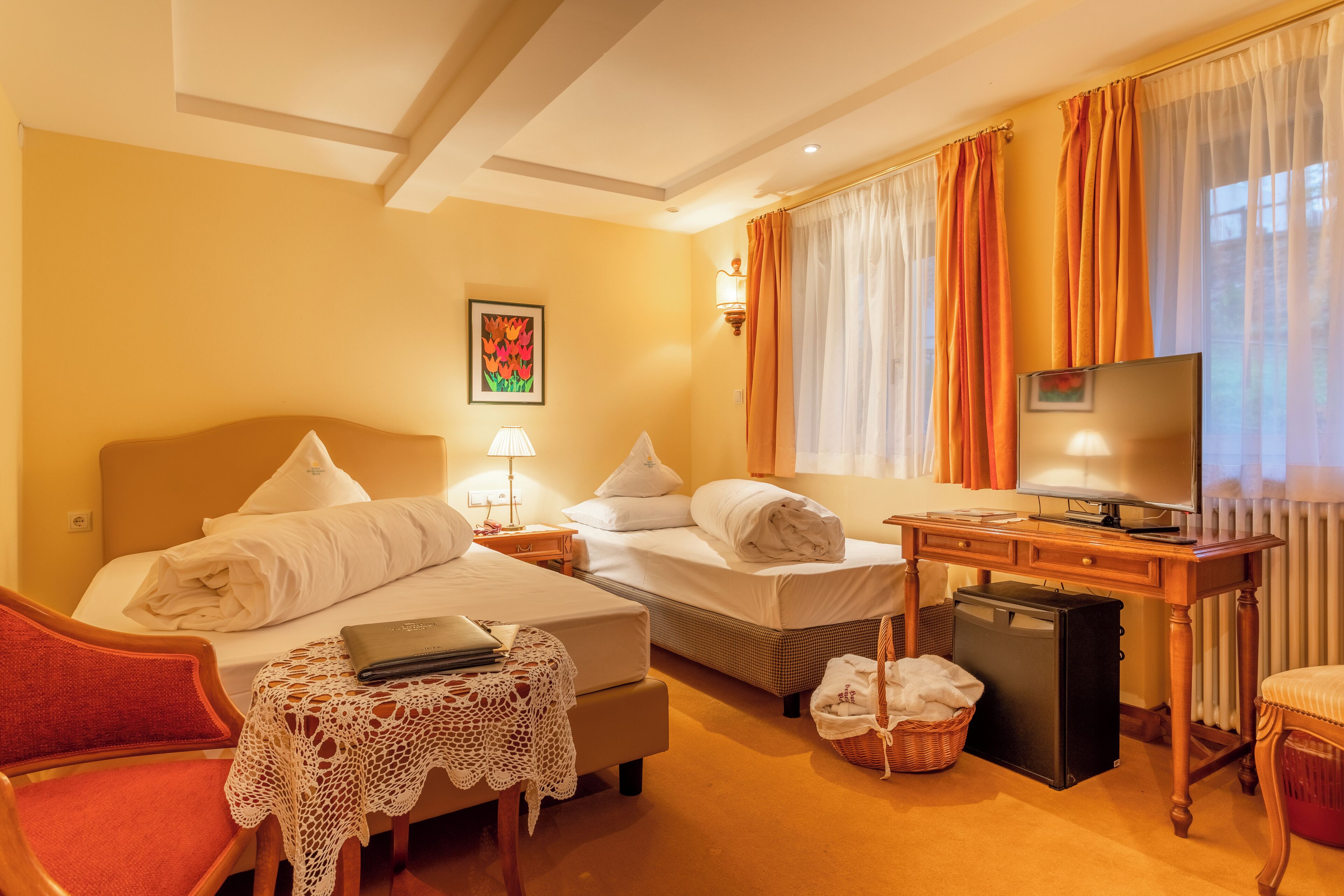 A hotel room with two beds, a desk with a television, a laptop, a small refrigerator, and a wicker basket.