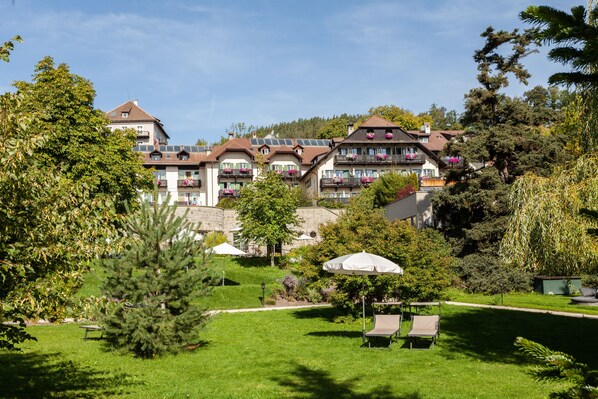 A hotel with multiple balconies, a garden area with a bench and umbrella, and a view of the surrounding landscape. - Bemelmans Post Hotel (Renon)
