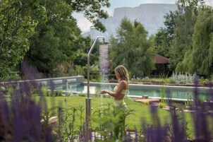 Una mujer de pie junto a una fuente en un jardín, con una piscina al fondo.