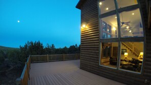 Une cabane en bois dotée d’une terrasse, de grandes fenêtres et d’un intérieur visible, avec un escalier et un coin salon.