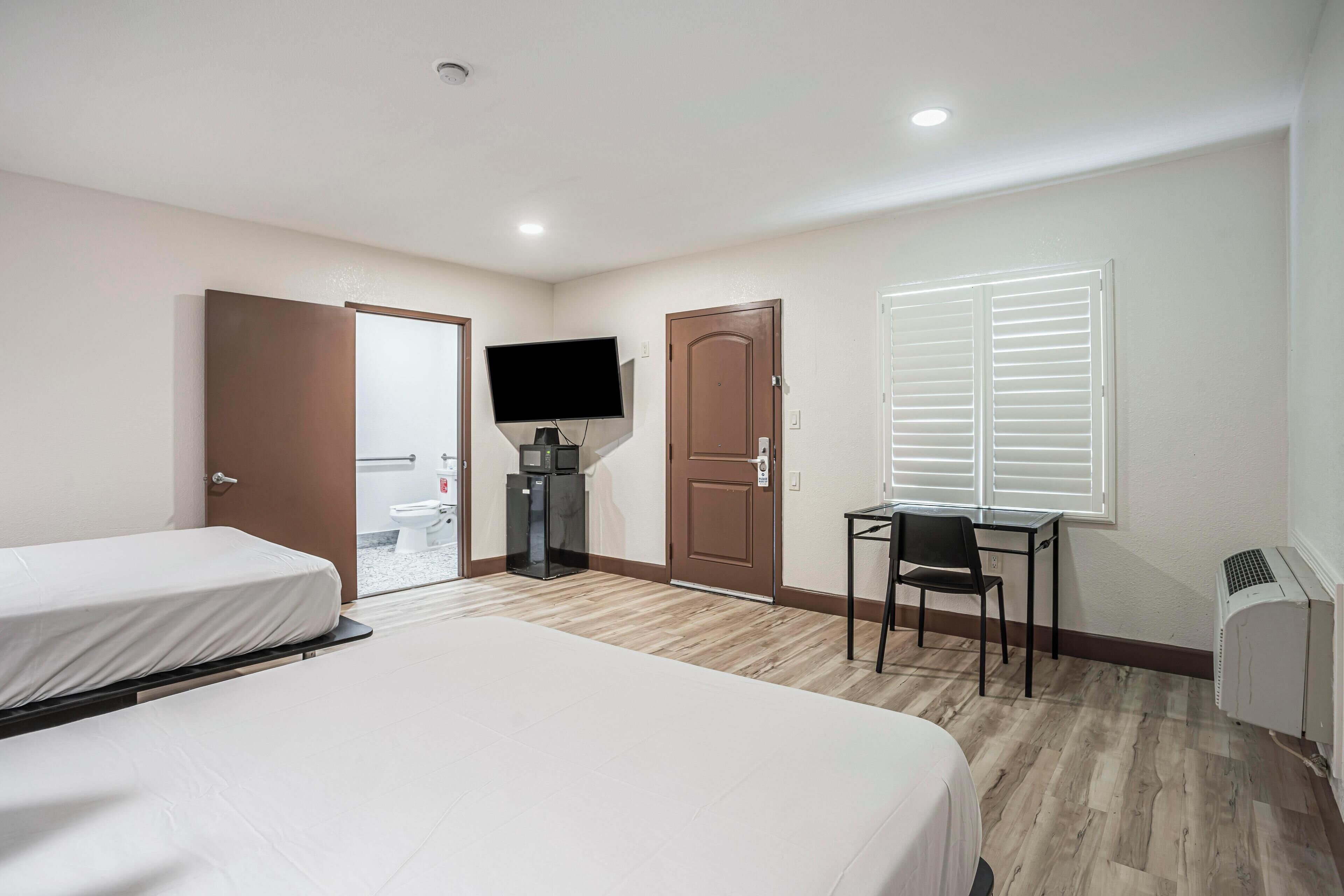 A hotel room with two beds, a TV, a desk with a chair, a window with blinds, and a door leading to a bathroom.