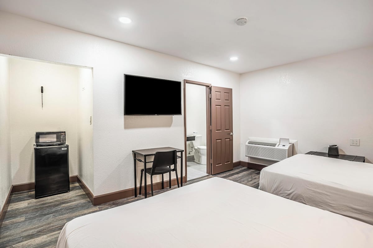 A hotel room with two beds, a desk, a microwave, a TV, and a bathroom visible through an open door.