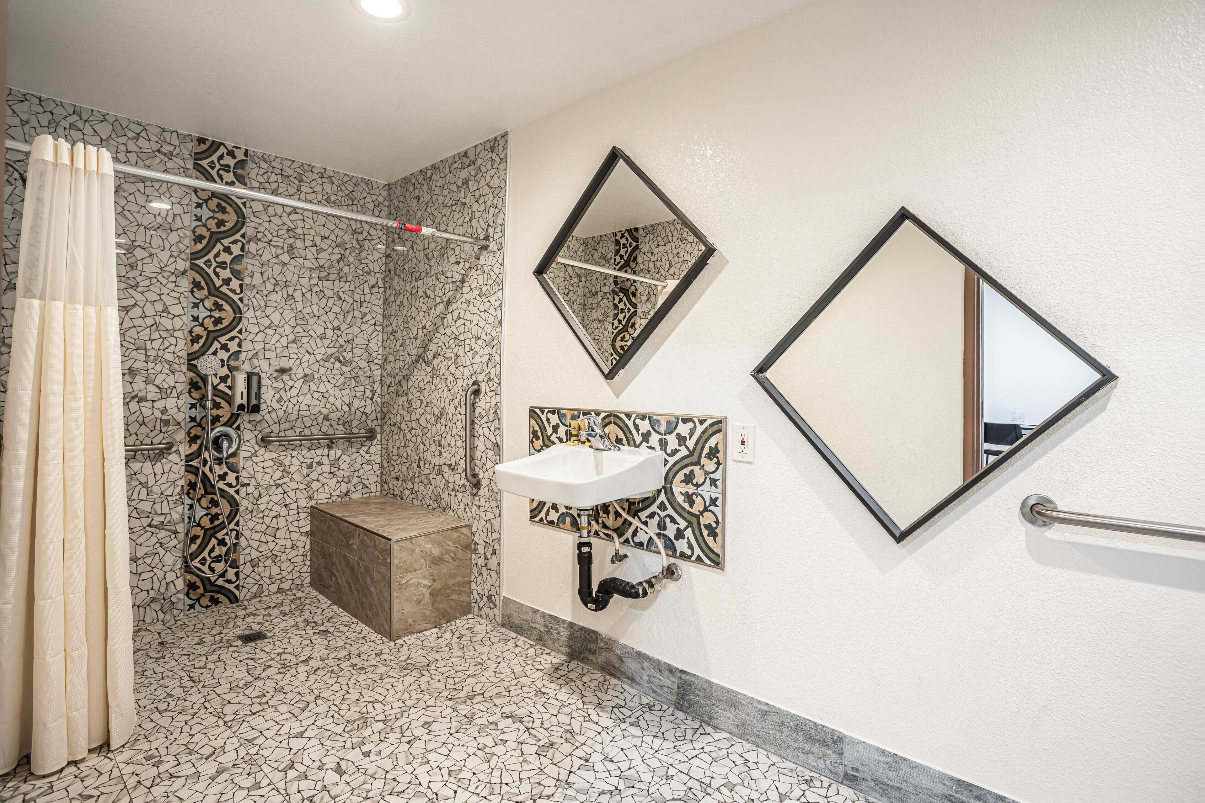 A bathroom with a mosaic floor, a shower with a curtain, a sink with a mirror, and a handrail.