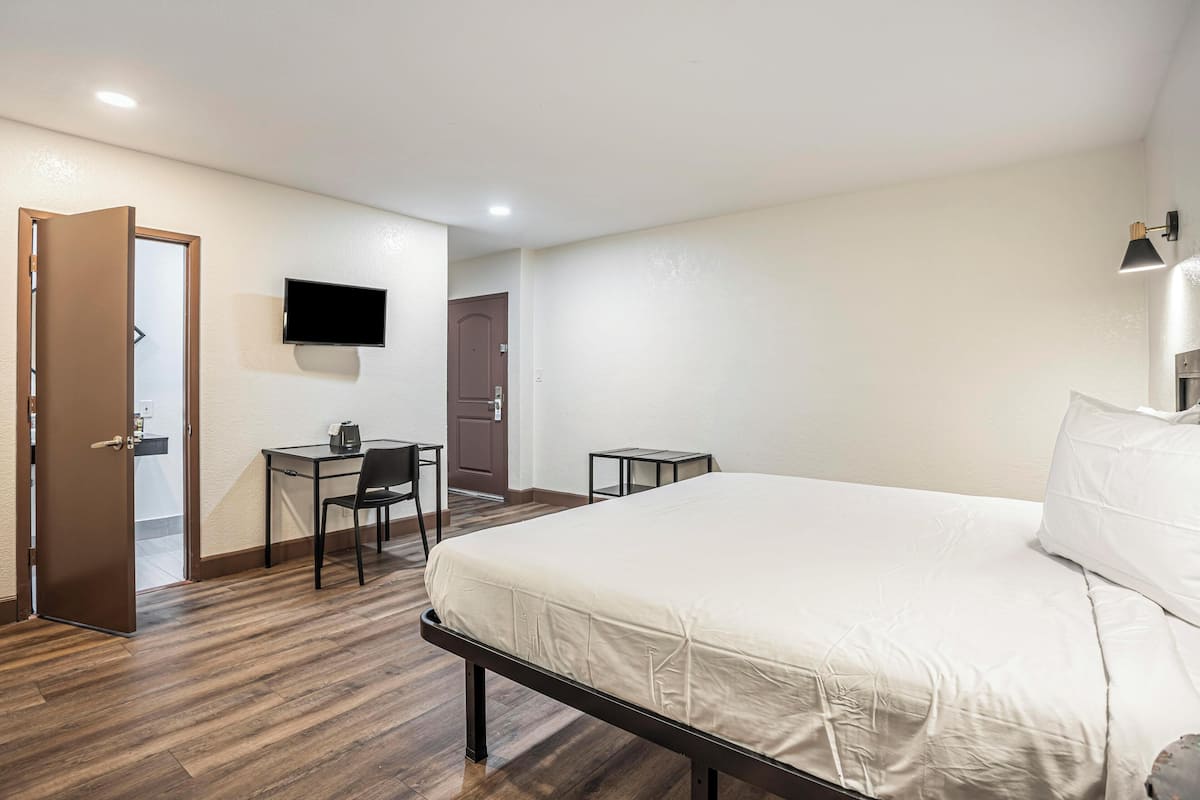 A hotel room with a bed, a desk with a chair, a television mounted on the wall, and a door leading to another room.