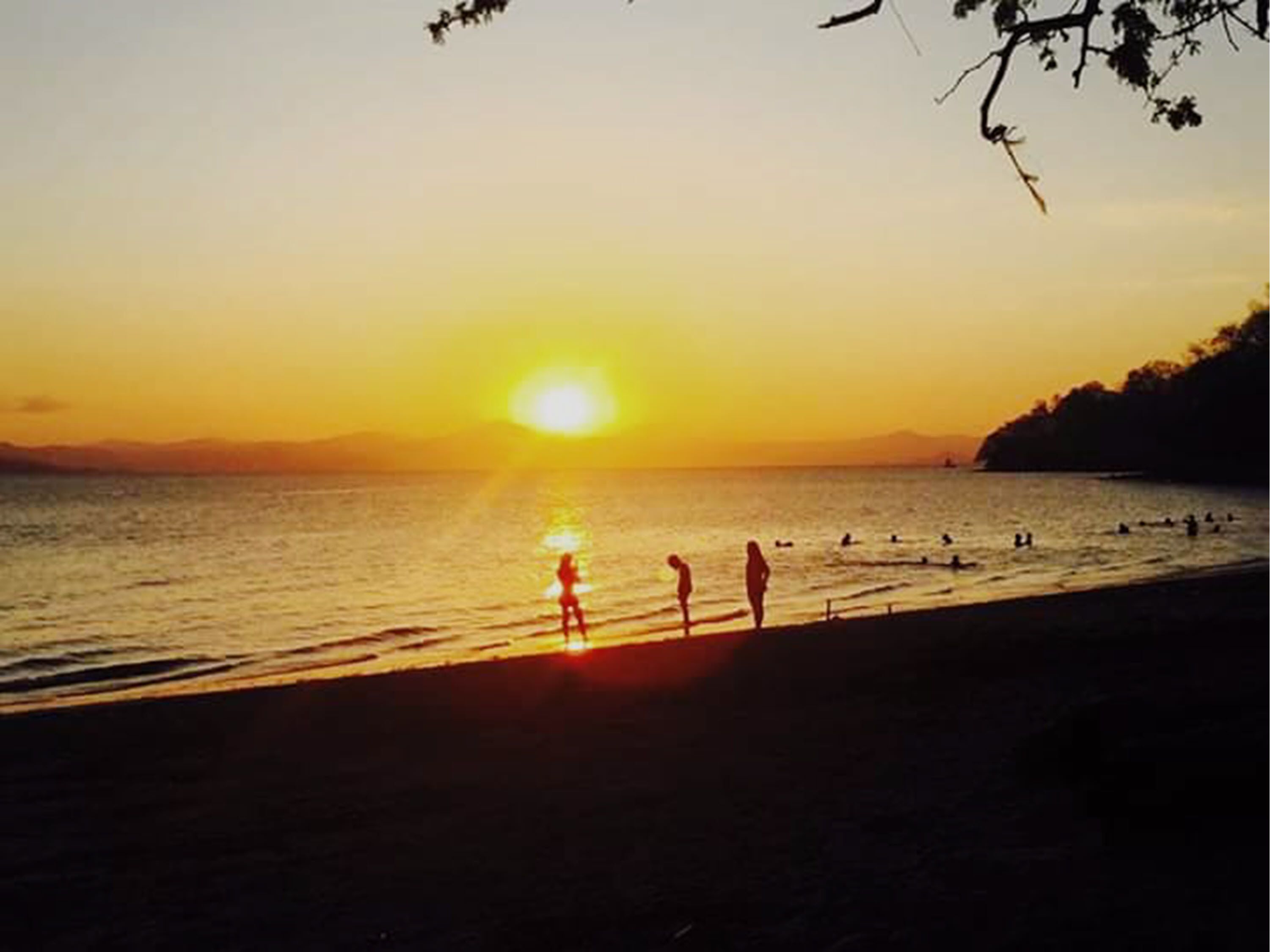 A beach scene with the sun setting over the water, silhouettes of people on the shore, and a distant mountain range.