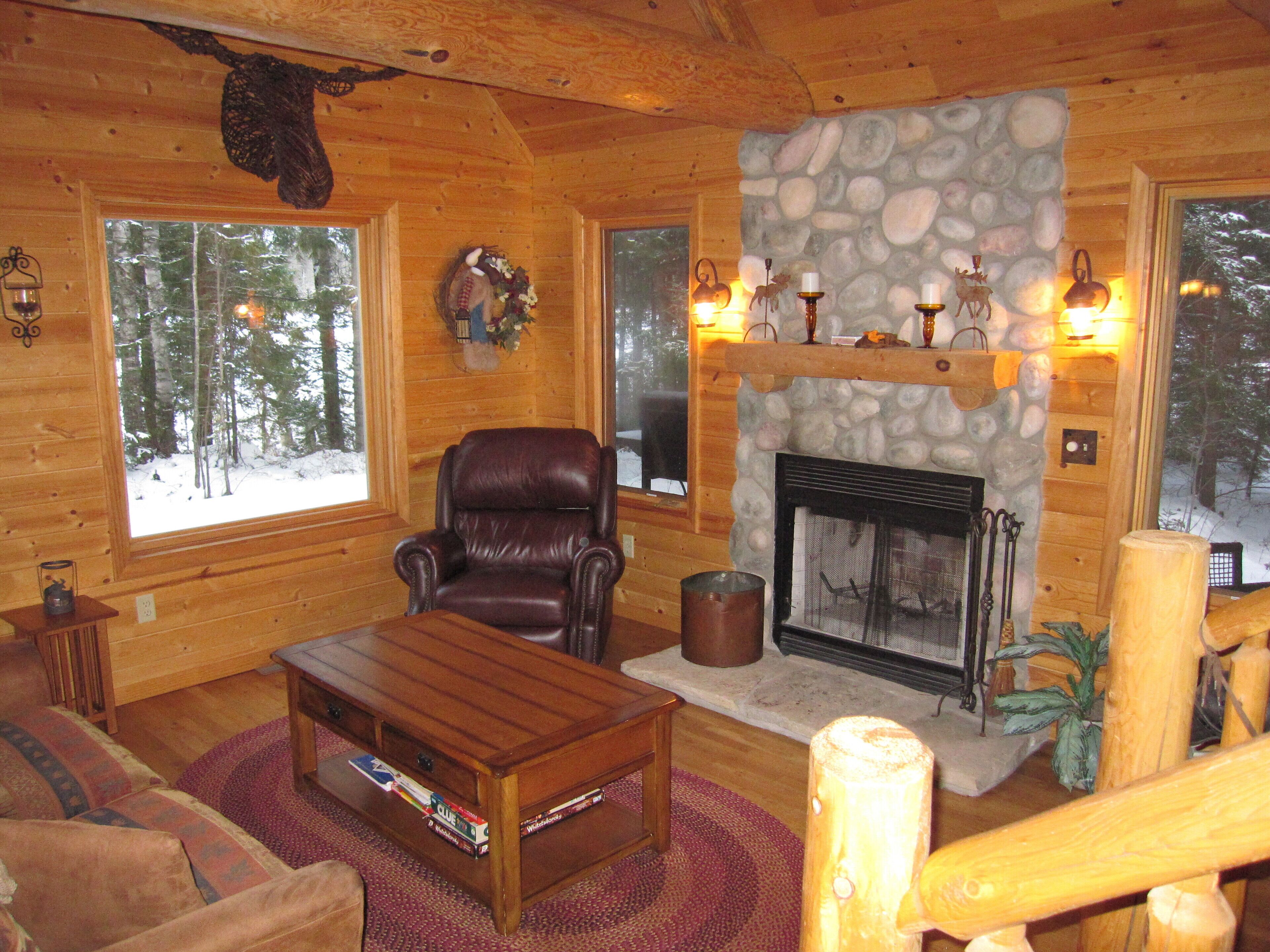 A cozy log cabin interior with a leather armchair, a fireplace, and large windows offering a view of snow-covered trees.
