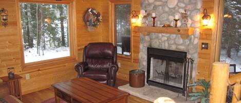 A cozy log cabin interior with a leather armchair, a fireplace, and large windows offering a view of snow-covered trees.